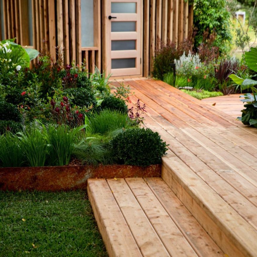Wooden deck with steps leading to a modern house entrance, surrounded by lush greenery and various plants. Vertical wooden slats partially cover the exterior, while Austral Bricks blend seamlessly into the natural aesthetic of the scene.