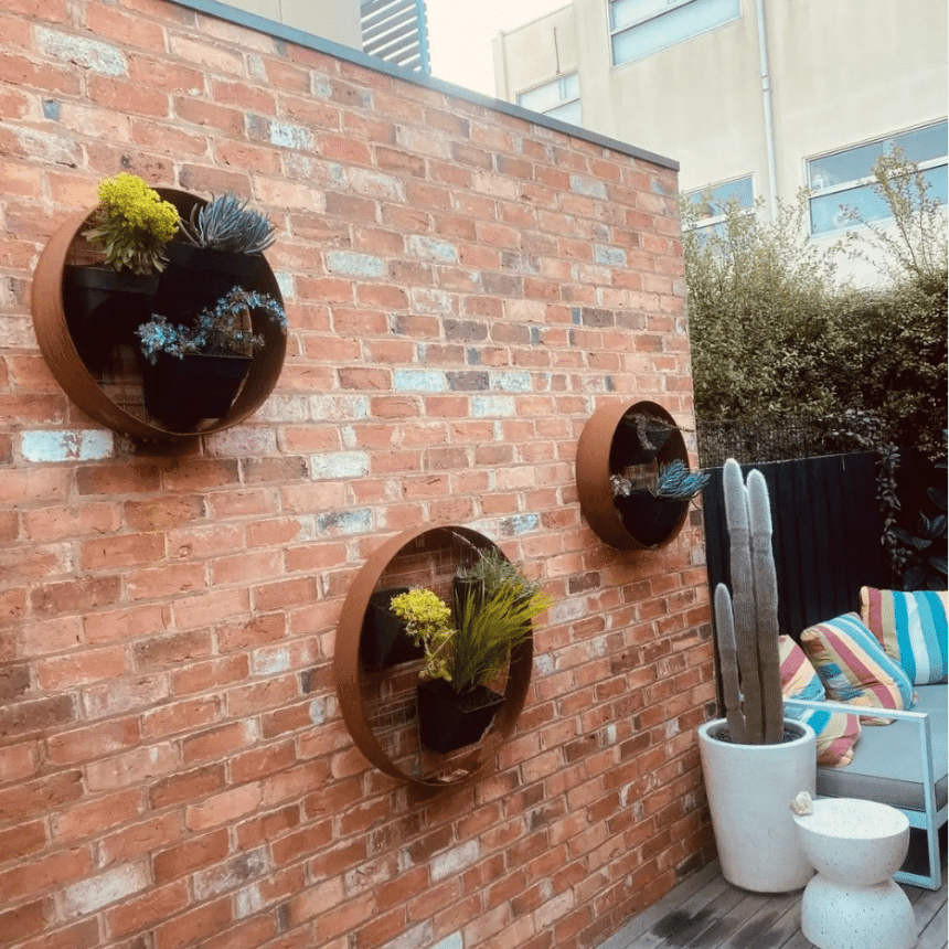 The outdoor patio showcases a striking brick wall adorned with three round, metal plant holders displaying various succulents. A cozy seating area with colourful cushions accompanies a tall, potted cactus and a white side table on the wooden deck.