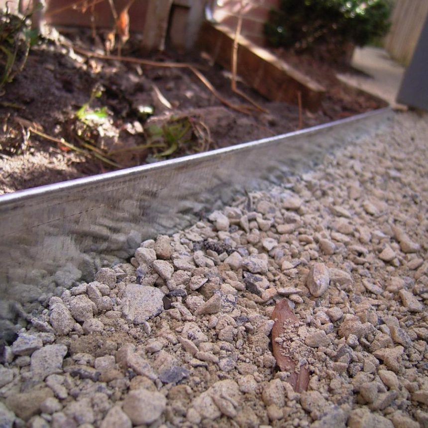 A close-up of a garden bed edge features metal edging that neatly separates gravel and soil. Small plants thrive against the backdrop of a sturdy Austral Bricks wall, adding a touch of timeless elegance to the scene.