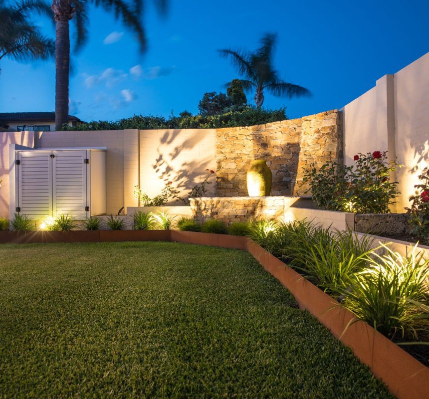 A well-lit backyard garden at dusk features lush green grass, a variety of plants, and a stone wall adorned with a decorative pot. Palm trees and other foliage stand out against the clear blue sky. Soft lighting enhances the landscape, beautifully complementing the timeless appeal of Austral Bricks.