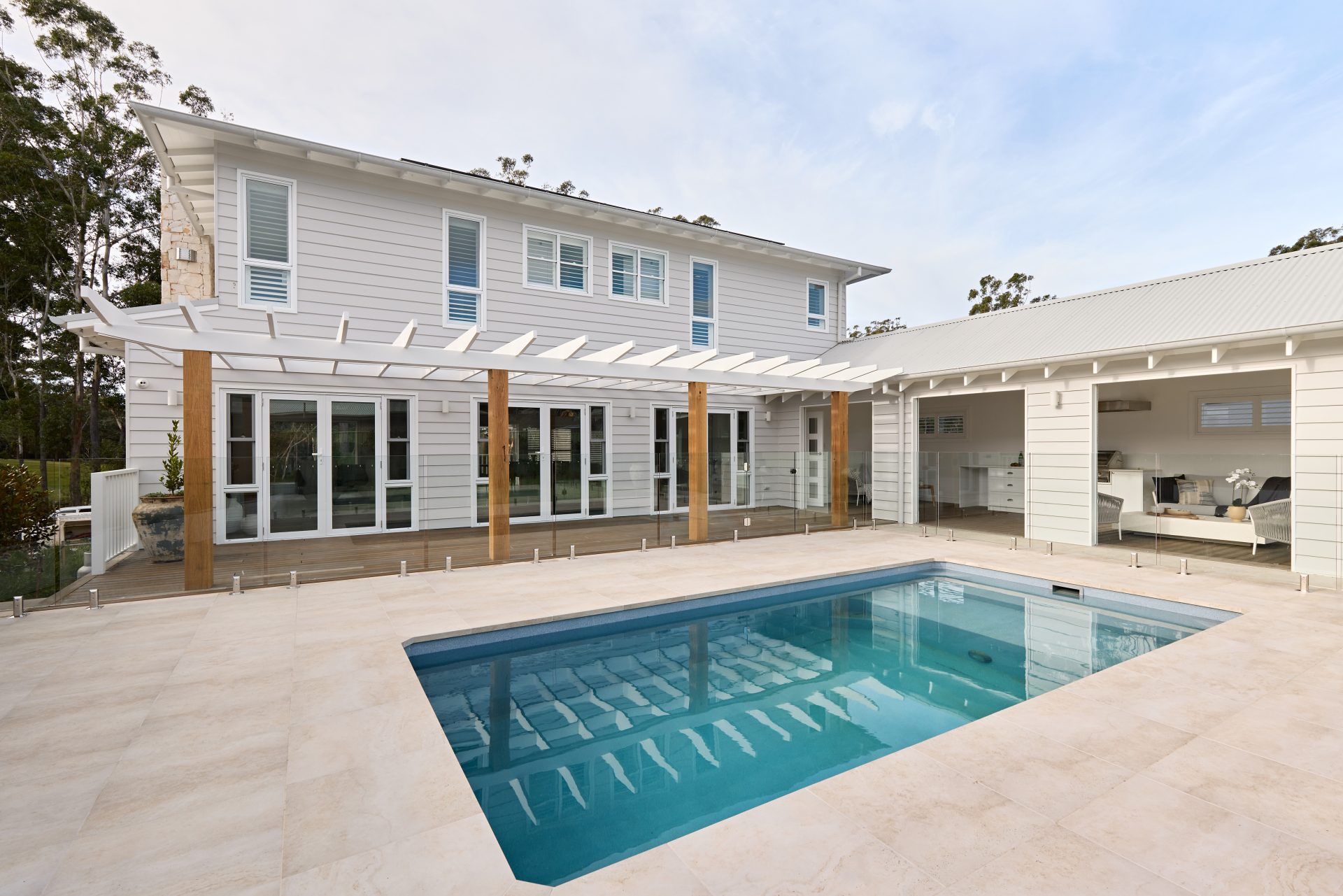 The outside of a house with a pool in the foreground paved with cream large format pavers. The house facade is white cladding with windows upstairs and downstairs overlooking the pool