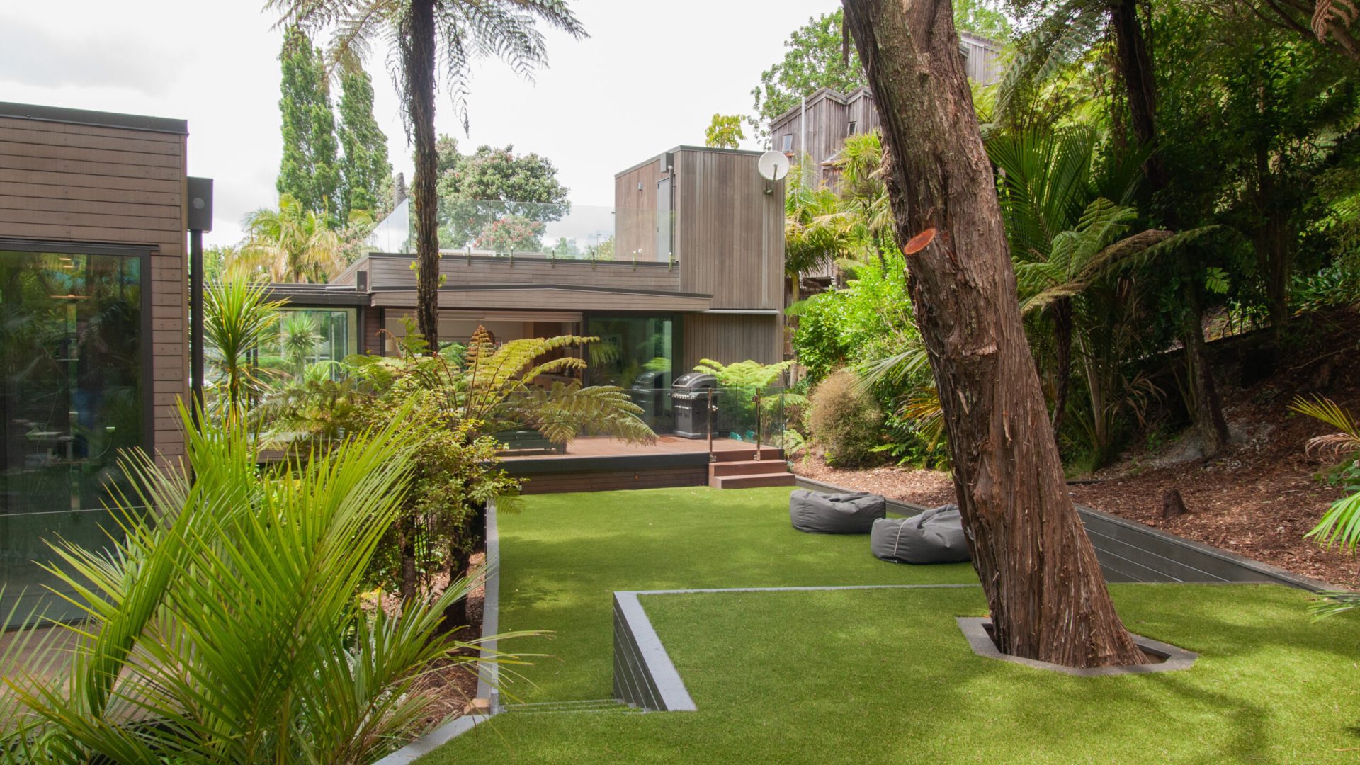 Modern house surrounded by lush greenery, featuring a glass and wood exterior. A Urban Turf Solutions Artificial Grass lawn area is dotted with a few plants and trees, with outdoor seating visible. The setting is serene and natural.
