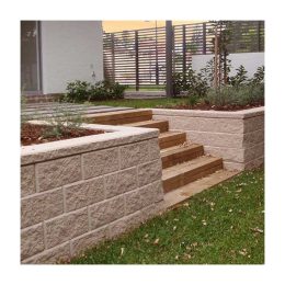 A garden pathway with wooden steps ascends through Austral Sydney Stone Block retaining wall , offering internal design inspiration. The area is landscaped with green grass and small shrubs, while a wooden slatted fence provides a charming backdrop.