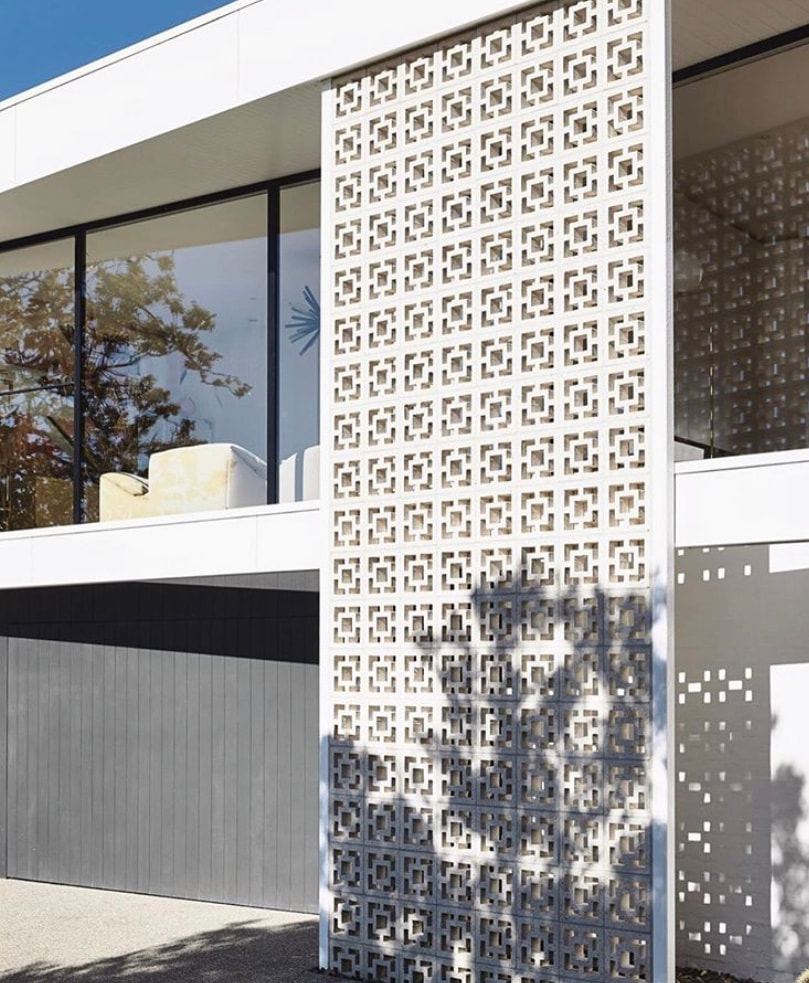 Modern building facade with a large, decorative panel featuring a Cubic Breeze blocks Double square cutouts. The panel casts intricate shadows on the building. Large windows reflect nearby trees and blue sky.