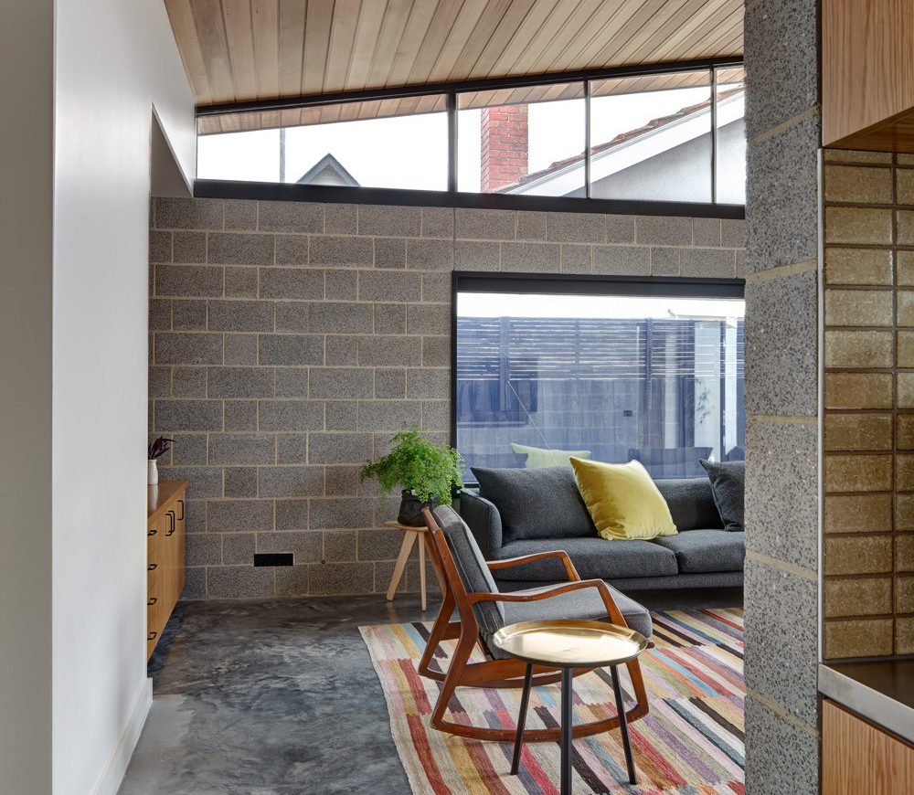 A modern living room exudes internal design inspiration with a concrete block wall and large window. A gray sofa adorned with green and yellow pillows rests on a colorful striped rug, beside a wooden chair with a white cushion. The wooden ceiling complements the light flooding the space.