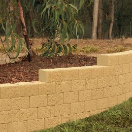 A curved, beige-colored retaining wall made of Baines Masonry stacked concrete blocks surrounds a landscaped area with mulch and trees. Behind the wall, tall trees rise against a forested backdrop, while the foreground features neatly trimmed green grass.