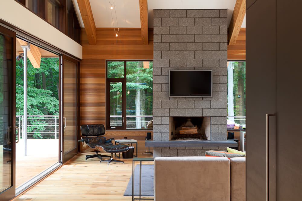 A modern living room showcases inspiring internal design, highlighted by a large stone fireplace with a mounted TV above. A leather lounge chair and ottoman sit nearby, while expansive windows provide views of lush trees outside. Wood-paneled walls add warmth to this stylish space.