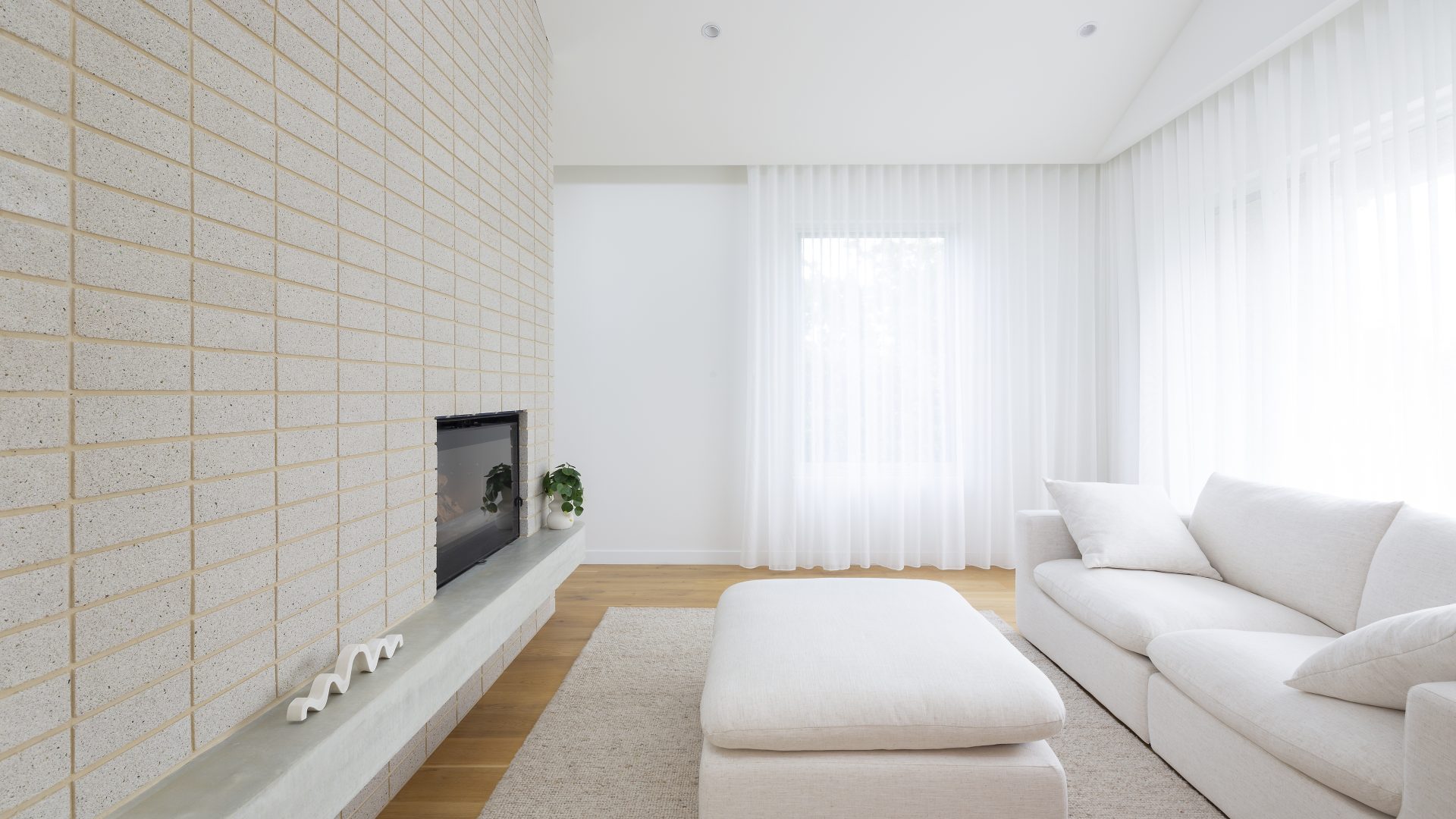 A minimalist living room with a white sofa, matching ottoman, and a beige textured rug crafted from sustainable building materials. The room features a modern fireplace set in a grid-patterned wall. Sheer white curtains cover the windows, allowing soft, natural light to fill the space.