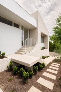 Modern white brick house with a minimalist design, constructed using sustainable building materials. It features a stairway with a sleek railing and a raised entrance. A path with rectangular stepping stones leads through a landscaped garden with small shrubs under a partly cloudy sky.