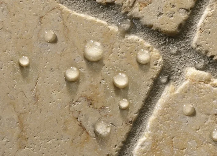 Close-up of a beige textured surface with irregular patterns, featuring droplets of water scattered across it. the surface appears porous, with distinct edges and variations in colour, adding an organic elegance.