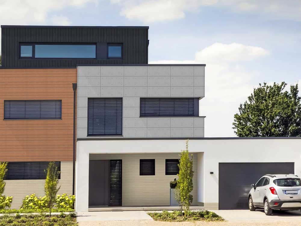 Modern two-story house with a mix of wooden, gray, and white exterior panels. A silver car is parked in the driveway beside the garage. Small trees and shrubs are planted in the front lawn under a partly cloudy sky.