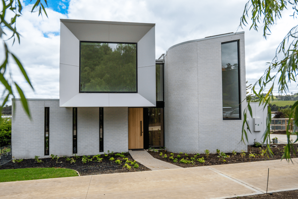 Modern two-story house with large square windows and curved walls, featuring a white brick exterior and minimalist design. Bricks line the pathway leading to the entrance, which is surrounded by a small landscaped garden.