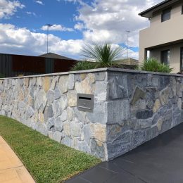 A stone retaining wall with a built-in mailbox surrounds a modern house. The sky is partly cloudy, and green plants adorn the top of the wall. A paved sidewalk and driveway lie in the foreground.