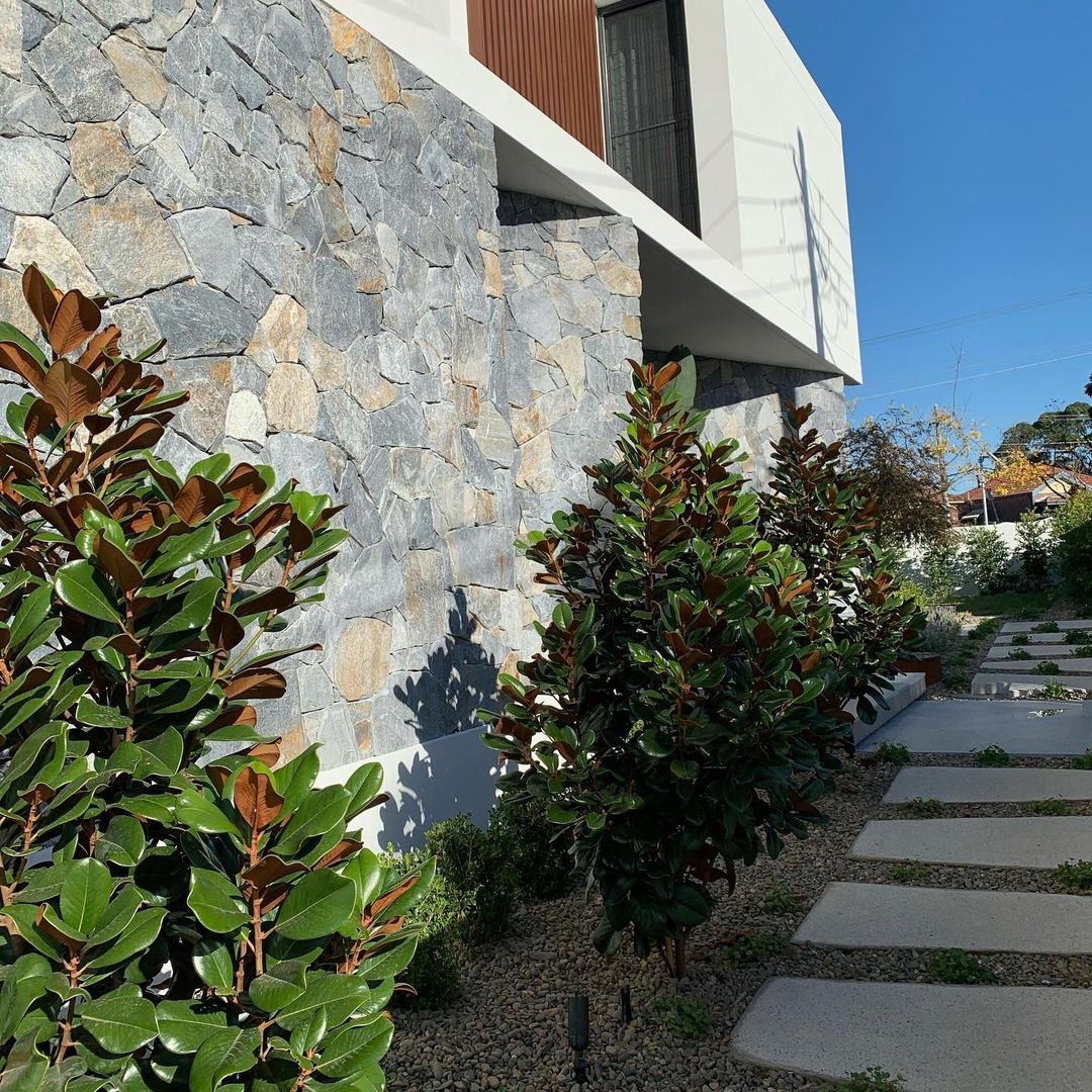 Modern house exterior with stone walls and large windows. The garden showcases retaining wall blocks seamlessly integrated with neatly arranged paving stones and lush green plants, all under a clear blue sky.