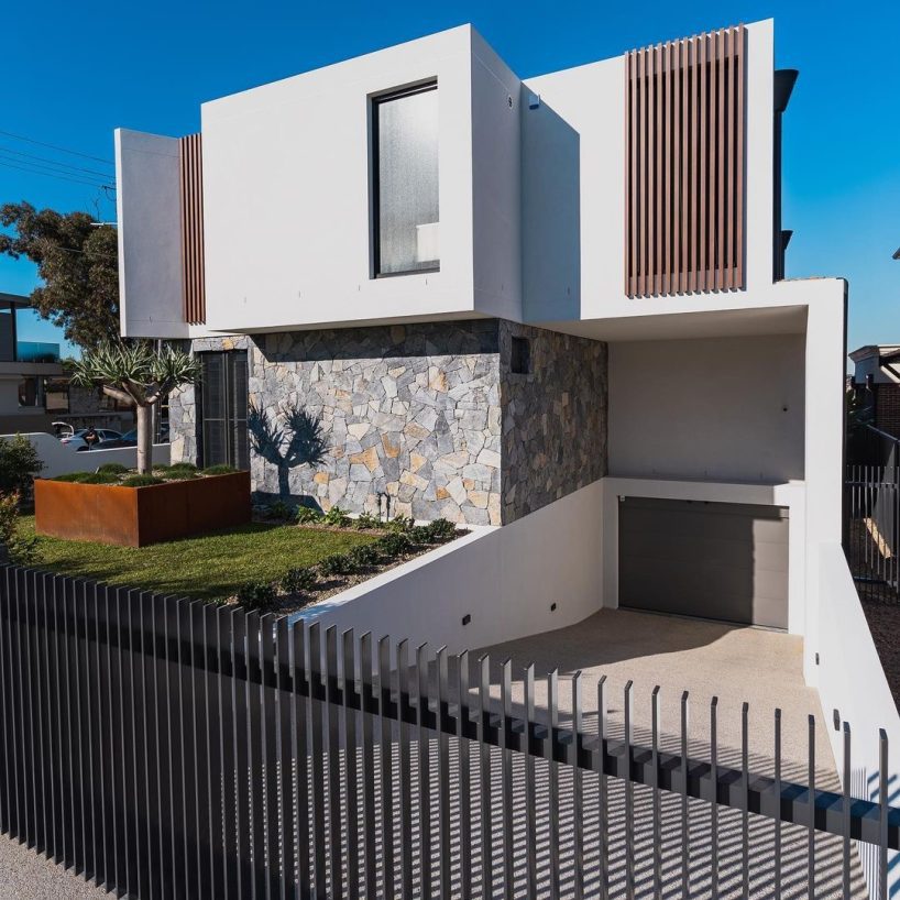 A modern two-story house with a stone and white facade, large window, vertical wooden slats, sloped driveway to a garage, and a landscaped front yard with green grass and plants enhanced by quality landscaping supplies.