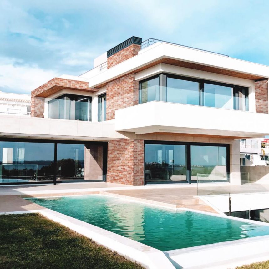 A modern two-story house with large glass windows and a mix of brick and white exterior. It features a terrace and a rectangular pool surrounded by grass, set against a clear blue sky.