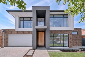 Modern two-story house with a mix of brick and gray exterior panels. Large windows, a wooden door, and a glass balcony are featured. The house is surrounded by a small lawn and trees, with a concrete driveway leading to a garage.