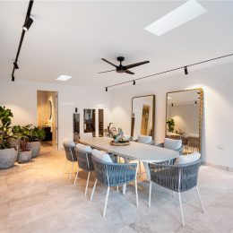 A modern dining room boasts a long table surrounded by wicker chairs, with large mirrors and lush potted plants adding a touch of elegance. Ceiling lights and a fan hang overhead, while landscaping supplies subtly accent the corner near the small bathroom.