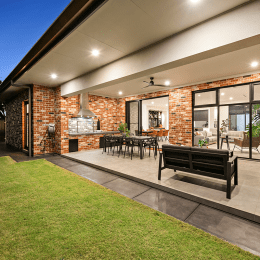 A modern outdoor patio with brick walls features a dining area, barbecue grill, and seating. Large sliding doors open to the interior, and the space is bordered by a lush green lawn. Warm lighting adds a cozy ambiance under a covered roof.