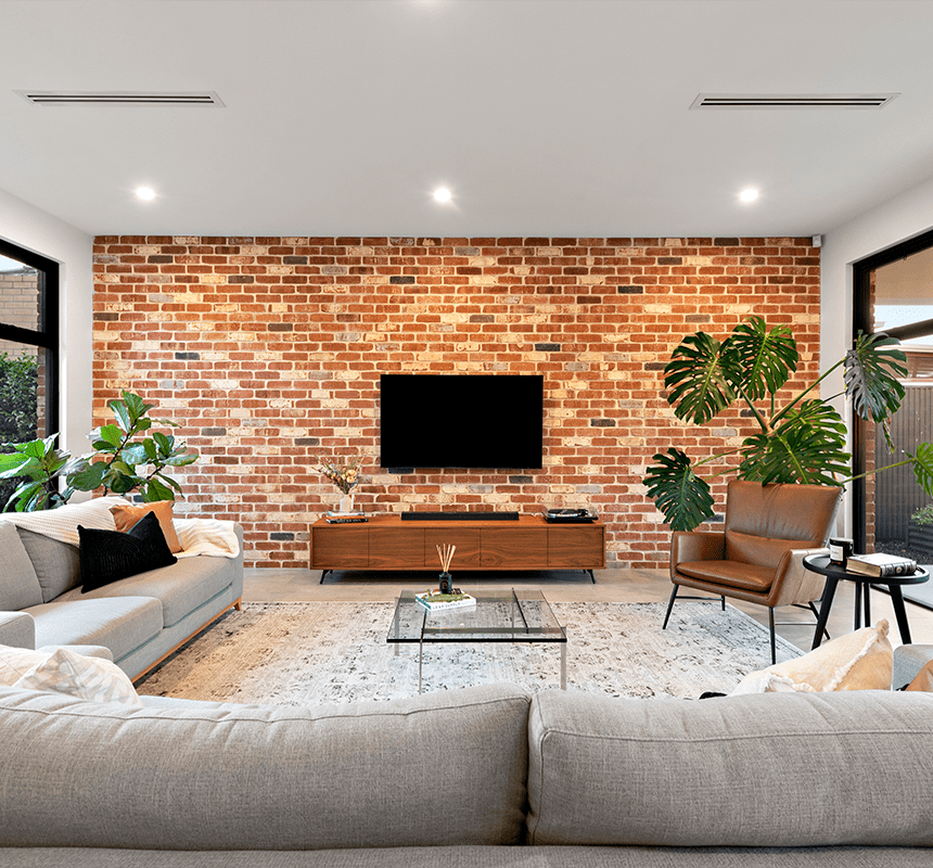 A modern living room with a brick accent wall features a flat-screen TV, light gray sofas, a white rug, a glass coffee table, two large potted plants, and a leather chair by a small side table. Large windows offer views of greenery outside.