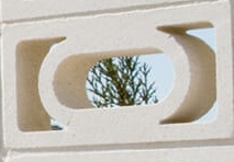Close-up of a rectangular coloured masonry block with a circular opening in the center. Through the opening, a small tree with green foliage is visible against a clear blue sky.