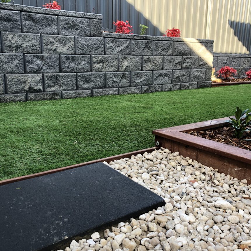 A landscaped backyard featuring neatly trimmed grass, a rock garden with white pebbles, and raised flower beds edged with timber. In the background, there are terraced stone walls with small red-leafed plants.