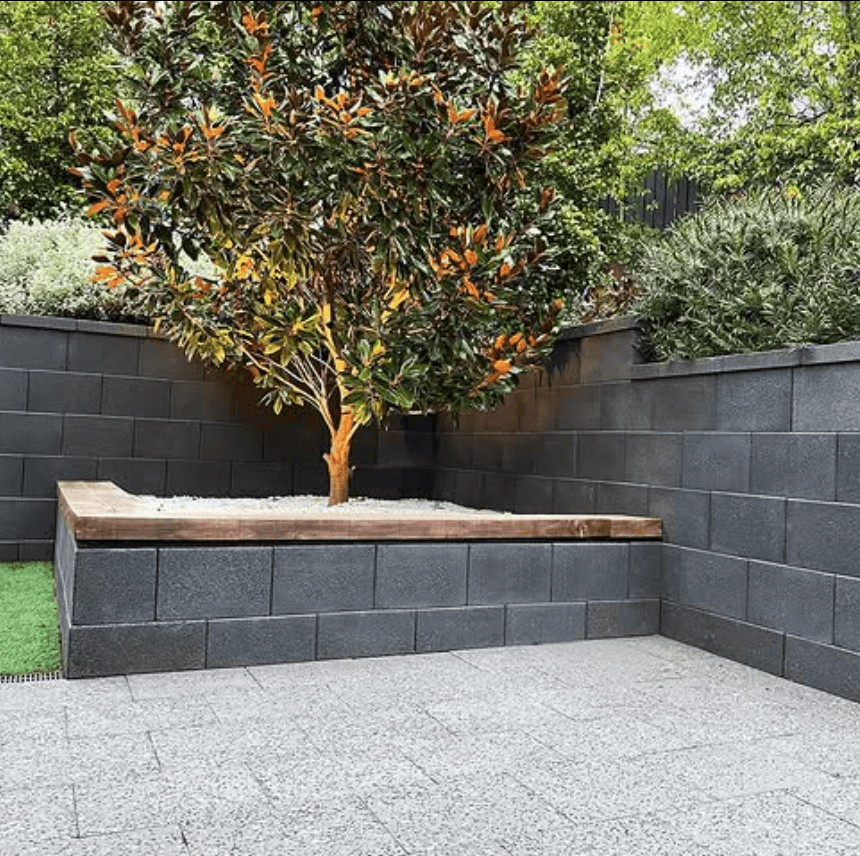 A landscaped backyard with a central raised planter bed containing a tree with foliage. The surrounding area features gray brick walls and a mix of greenery. The ground is paved with stone tiles and there is a strip of artificial grass.