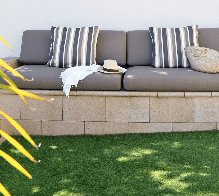 Outdoor seating area with a beige cushioned bench and striped pillows against a white wall. A straw hat and light blanket rest on the bench. Nearby, a plants leaves are visible, and the space features a grass lawn.