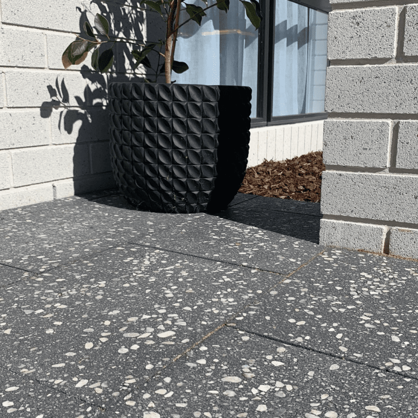 A modern patio with dark terrazzo floor tiles featuring white speckles. A black textured planter holds a plant, set against a white brick wall with a window. Mulched garden bed is visible in the background.