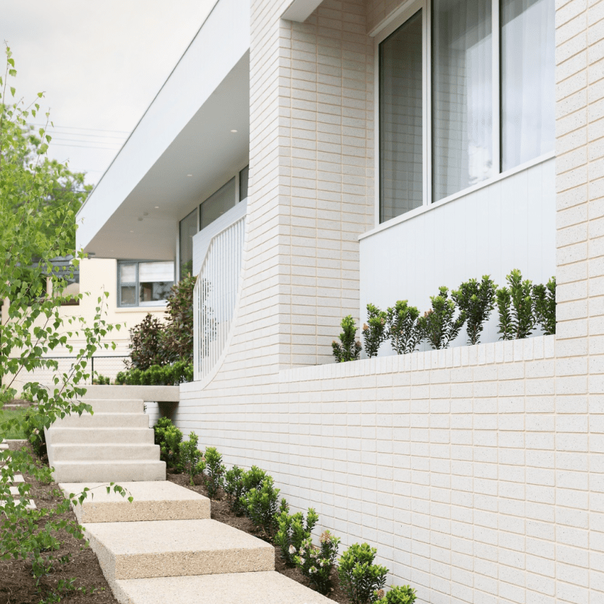 Modern white brick building with large windows and a curved wall. A stepped pathway leads up, surrounded by small shrubs and greenery, under an overcast sky.