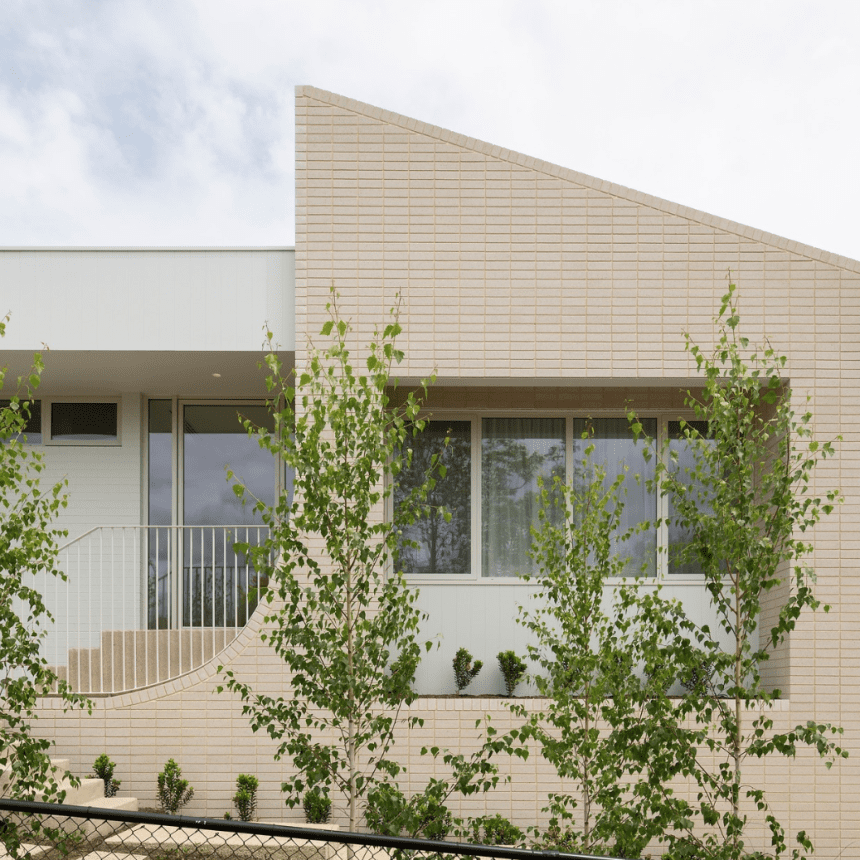 A modern building with a light beige brick exterior, featuring large windows and minimalist design. The structure includes a distinctive sloped roof and a white metal railing. Young trees are planted along the facade, adding greenery to the scene.