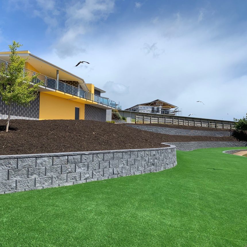 A modern building with yellow accents stands on a hillside. The foreground features a manicured lawn and stone retaining walls. Several birds fly in the clear blue sky. A small tree is planted on the slope.