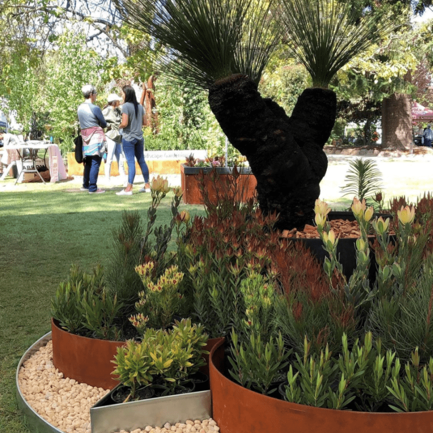 A garden scene features circular planter beds bordered by FormBoss Garden Edging, filled with green and red plants. A prominent large grass tree with a dark trunk is visible. People are in the background under a canopy, surrounded by grass and trees.