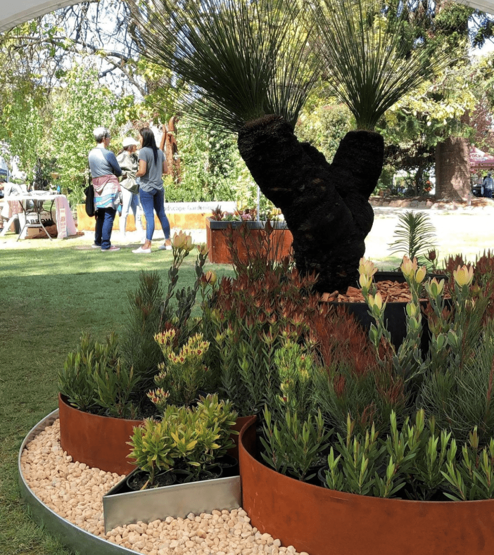 A garden scene features circular planter beds bordered by FormBoss Garden Edging, filled with green and red plants. A prominent large grass tree with a dark trunk is visible. People are in the background under a canopy, surrounded by grass and trees.
