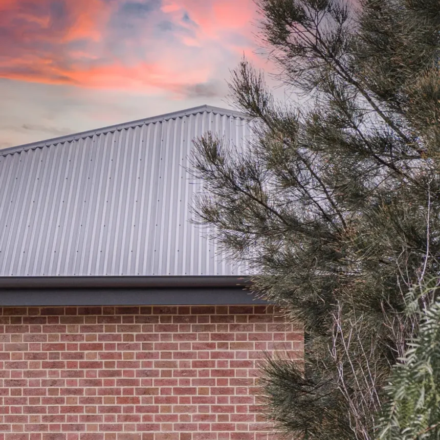 Littlehampton Bricks and Pavers Light Cabernet Bricks in a wall of a house in a close up photo of half a wall and the triangle grey tin roof with a pink sunset in the background and an Australian native tree in front of the wall
