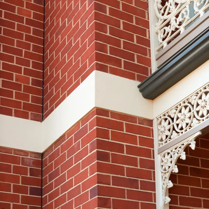 A close-up of a red brick building corner, featuring Original Adelaide Red Bricks with a horizontal cream-colored stripe and ornate white wrought iron balcony railings.