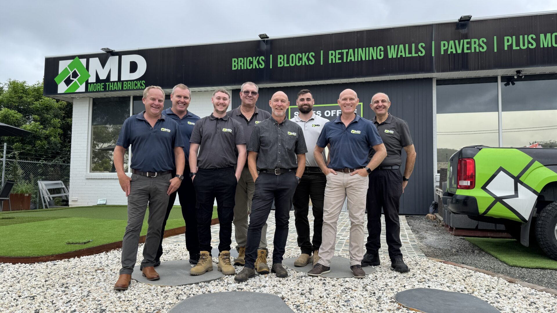 Eight men wearing branded shirts stand and smile outside a building with an "MD More Than Bricks" sign. A branded vehicle is parked on the right, beside landscaping with rocks and artificial grass. Learn more about us and our team spirit.