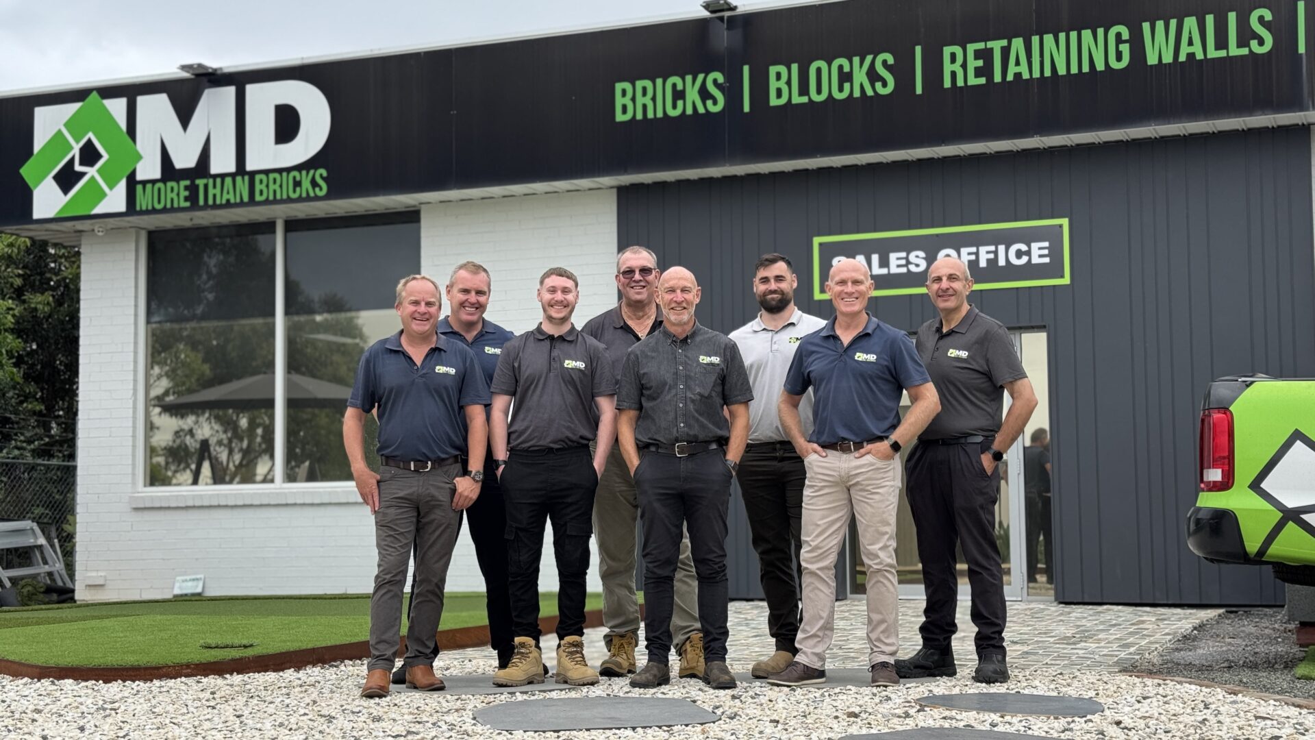 Eight men stand smiling in front of a building with a sign reading MD More Than Bricks, Bricks | Blocks | Retaining Walls, next to a sales office and a vehicle with the company logo, where Helpful Videos are also offered for customers.