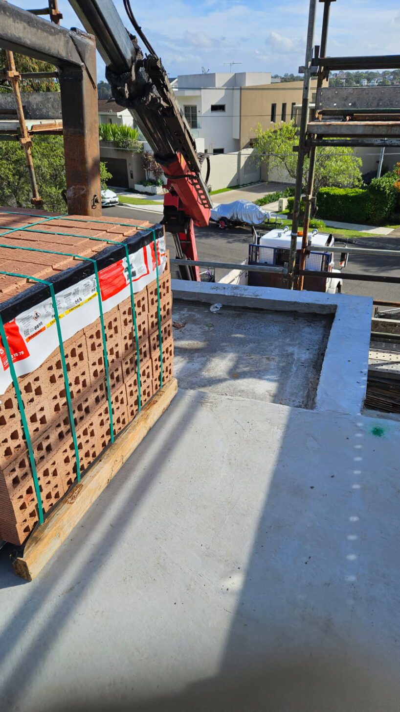 A pallet of bricks, prepared for brick matching, is being lifted by a crane onto a concrete rooftop at a construction site, with a street, cars, trees, and modern buildings visible in the background.