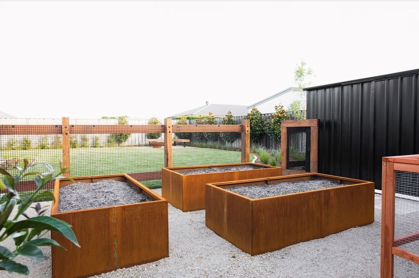 Three large rectangular metal raised garden beds filled with soil are arranged on a gravel surface in a fenced backyard, with a black shed and a grassy lawn in the background.