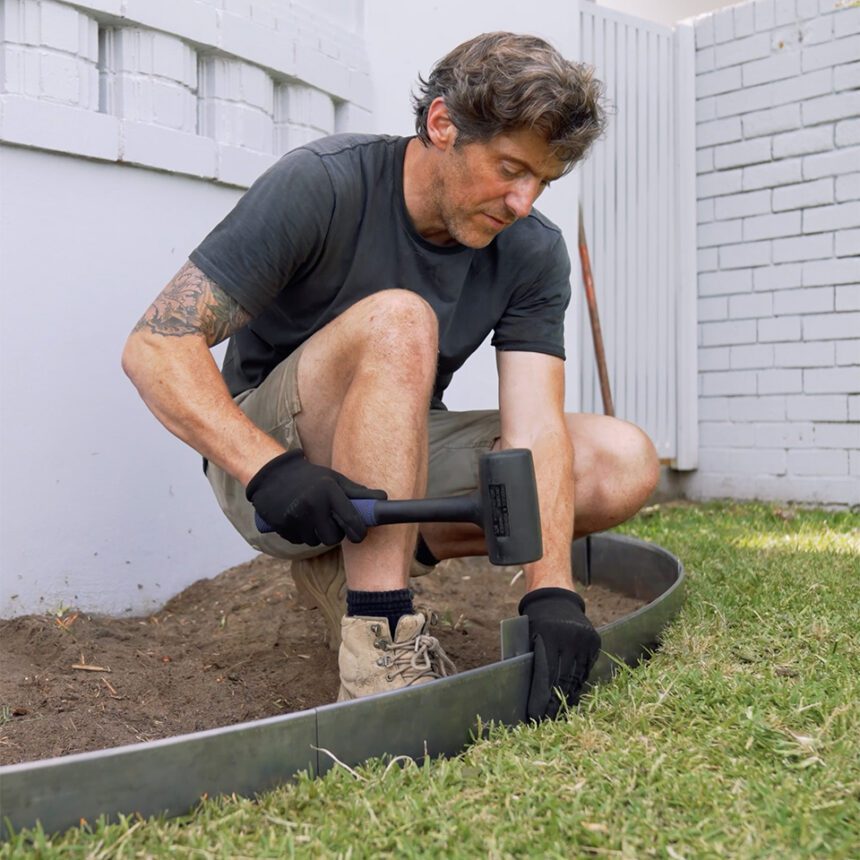 A man wearing gloves uses a rubber mallet to secure metal edging between a garden bed and grass in a backyard, kneeling on one knee.