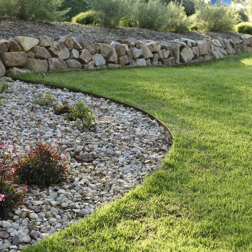 A landscaped garden with a curved border separates a gravel bed with plants from a neatly mowed green lawn. In the background, there is a low stone retaining wall and more greenery.