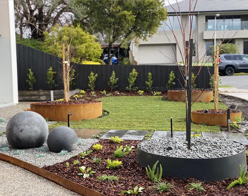 Modern landscaped front yard with circular garden beds, metal edging, gravel, decorative concrete spheres, young trees, mulch, and neatly trimmed grass, set in a suburban neighborhood with houses and parked cars in the background.