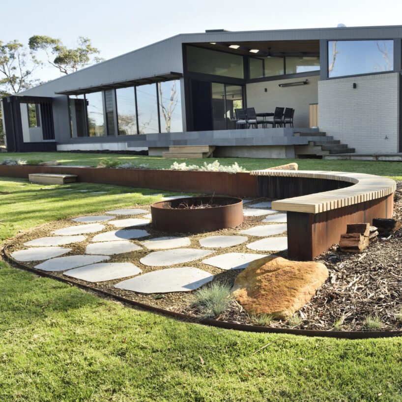 A modern house with large windows overlooks a landscaped backyard featuring FormBoss Metal Edging, a circular fire pit, stone pavers, curved benches, and neatly trimmed grass. Trees and blue sky are visible in the background.