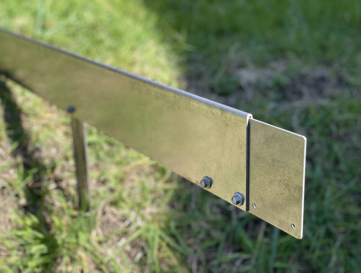 A close-up of a metal bracket with bolts securing two overlapping plates, standing upright on grassy ground in bright sunlight.