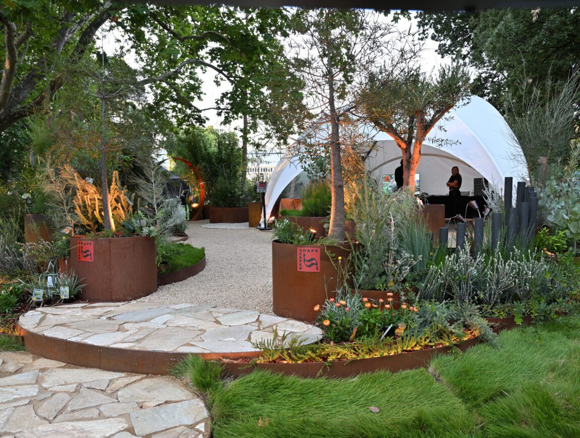 A landscaped garden with stone and gravel paths, lush plants, metal planters, and a white dome tent in the background under large trees. A person stands inside the tent.
