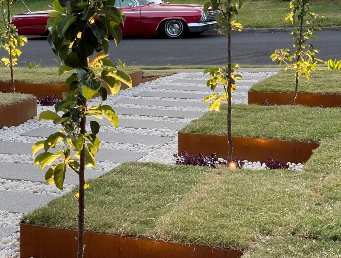 Young trees planted in square beds with white stones and small purple plants line a neat garden path. Soft lighting highlights the landscaping. A vintage red car is parked on the street in the background.