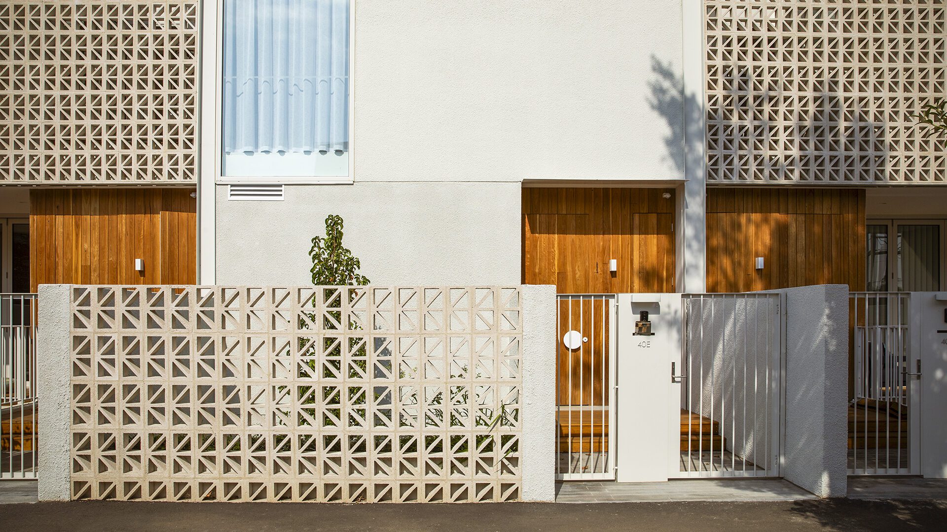Modern residential building with a geometric-patterned breeze block fence, wooden doors, and light-colored walls. A small tree grows inside the fenced area, and sunlight casts shadows on the facade.