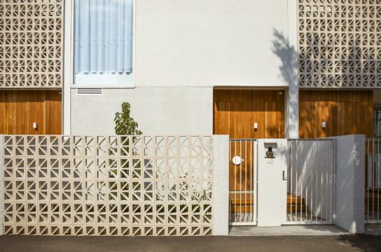 Modern residential building with a geometric-patterned breeze block fence, wooden doors, and light-colored walls. A small tree grows inside the fenced area, and sunlight casts shadows on the facade.