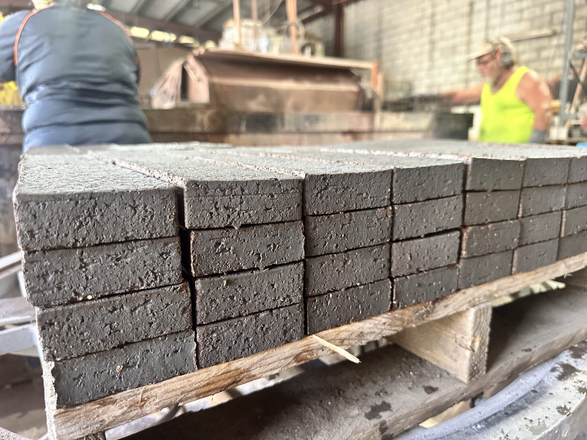 A close-up view of neatly stacked, dark recycled bricks on a wooden pallet in an industrial workshop, with two people working in the blurred background. They are cutting face bricks into brick slips for a lightweight brick cladding system.